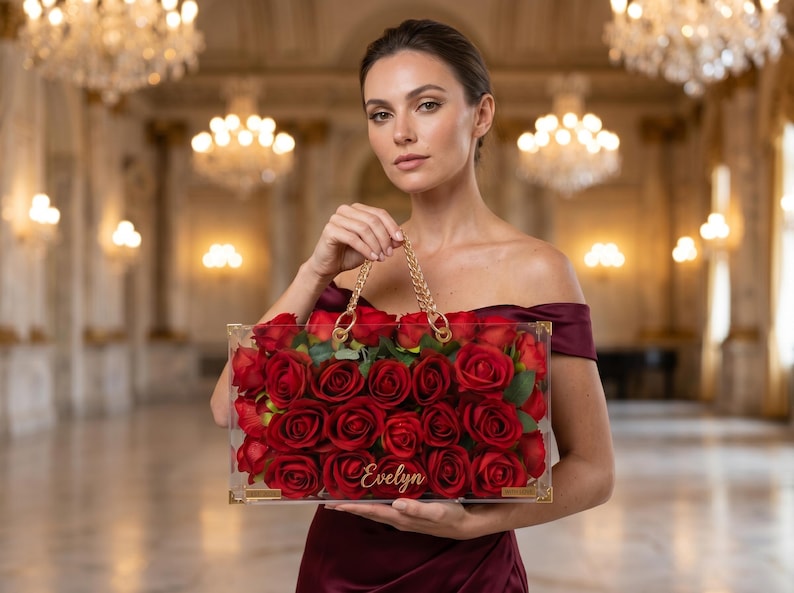 May include: A clear rectangular handbag with gold trim and a gold chain handle, filled with vibrant red roses. The name "Evelyn" is inscribed on the front. The bag is held against a blurred background of a grand hall.