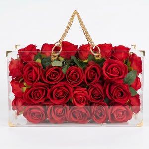 May include: A clear acrylic handbag filled with vibrant red roses. The bag features gold-colored corner accents and a gold chain handle. The roses are in full bloom, creating a striking visual contrast against the transparent bag.