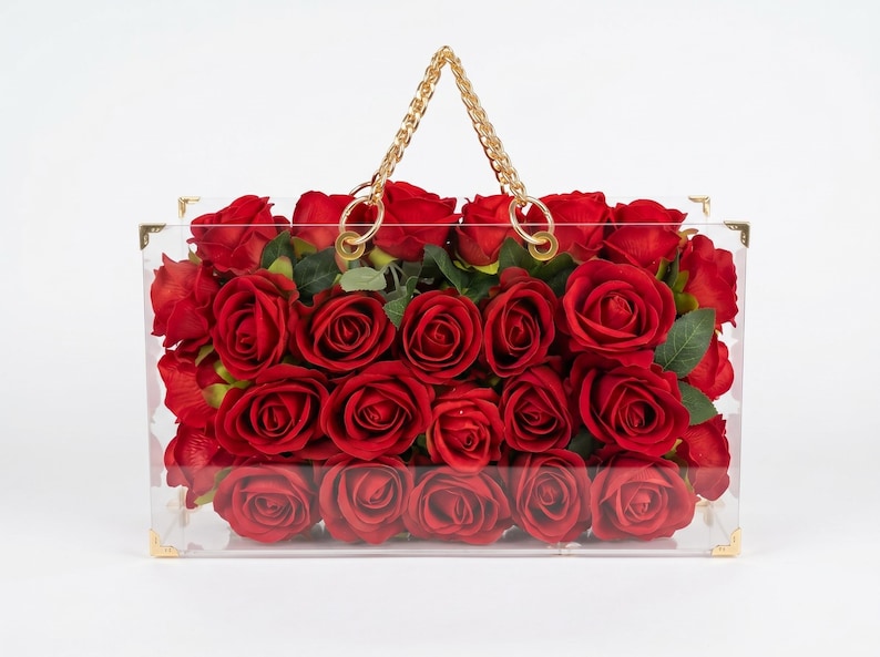 May include: A clear acrylic handbag filled with vibrant red roses. The bag features gold-colored corner accents and a gold chain handle. The roses are in full bloom, creating a striking visual contrast against the transparent bag.