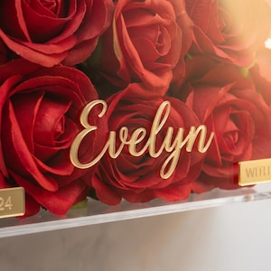 May include: A clear acrylic box filled with vibrant red roses. The name "Evelyn" is displayed in gold script across the front. Gold accents include corner pieces and a small plaque that reads "WITH LOVE".