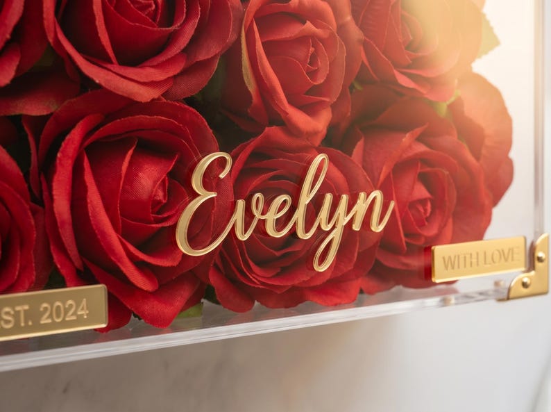 May include: A clear acrylic box filled with vibrant red roses. The name "Evelyn" is displayed in gold script across the front. Gold accents include corner pieces and a small plaque that reads "WITH LOVE".