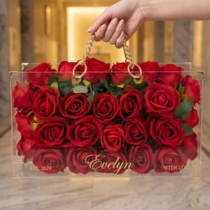 May include: A clear acrylic box filled with red roses, featuring gold-colored hardware and a chain handle. The box is personalized with the name "Evelyn" and the text "EST. 2024 WITH LOVE".