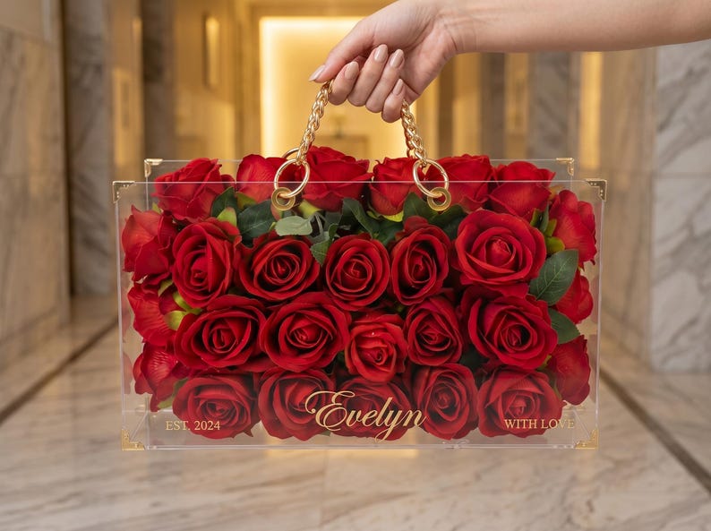 May include: A clear acrylic box filled with red roses, featuring gold-colored hardware and a chain handle. The box is personalized with the name "Evelyn" and the text "EST. 2024 WITH LOVE".