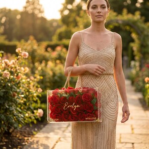 May include: A clear acrylic handbag filled with red roses, featuring a gold chain strap. The bag is personalized with the name "Evelyn" and the words "WITH LOVE". The background shows a garden with rose bushes and a woman in a beaded dress.