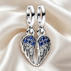 May include: A pair of silver earrings with a wing design. Each wing is decorated with blue and clear gemstones. The earrings are suspended from a circular hoop. The background is a white, silky fabric.