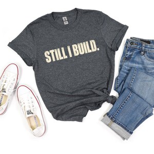 Könnte beinhalten: Ein dunkel anthrazitfarbenes Kurzarm-T-Shirt mit dem Text STILL I BUILD. in verwaschener cremefarbener Schrift auf der Brust, kombiniert mit einer blauen Jeanshose und weißen Canvas-Sneakern mit niedrigem Schaft und rot-blauen Details.