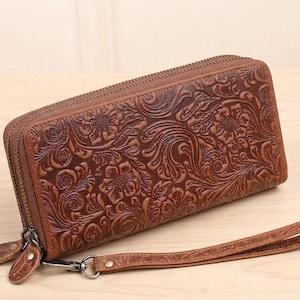 May include: A brown leather wallet with a floral embossed design. The wallet has a zipper closure and a wrist strap. The leather is a warm brown color, and the floral pattern is detailed and intricate. The wallet is rectangular in shape.