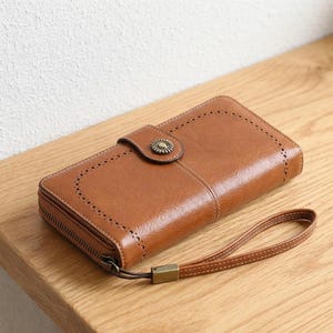 Könnte beinhalten: Eine braune Ledergeldbörse mit einer Handschlaufe. Die Geldbörse hat einen dekorativen Knopfverschluss und einen Reißverschluss. Das Leder hat ein strukturiertes Aussehen mit dekorativen Nähten an den Kanten. Die Geldbörse ist rechteckig.