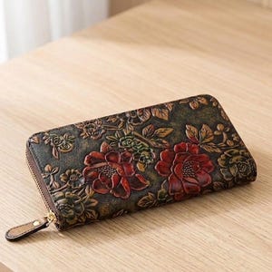 Puede incluir: Cartera de cuero marrón oscuro con diseño floral en relieve. La cartera presenta flores en relieve en rojo, verde y marrón, con cierre de cremallera dorada. La cartera rectangular parece ser de cuero de alta calidad.