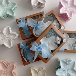 Op de afbeelding: Een verzameling zeestervormige keramische schaaltjes in pasteltinten roze, blauw en wit, gerangschikt rond rechthoekige geschenkdoosjes van kraftpapier met doorzichtige vensters, blauwe strikken en kleine witte snoepjes op blauw vulmateriaal.