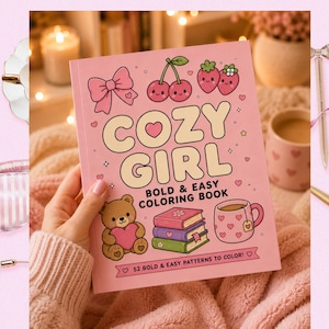 May include: A pink coloring book titled "COZY GIRL" with illustrations of a bow, cherries, strawberries, a teddy bear, books, and a mug. The book is held in a person's hand. Other items in the image include a sleep mask, a pen, and a decorative bird.