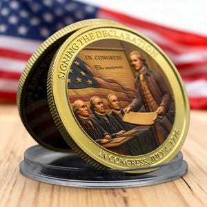 Könnte beinhalten: Eine goldfarbene Gedenkmünze mit einer Darstellung der Unterzeichnung der Unabhängigkeitserklärung. Die Münze trägt die Aufschrift "Signing the Declaration" und "In Congress, July 4, 1776". Im Hintergrund ist eine amerikanische Flagge zu sehen.