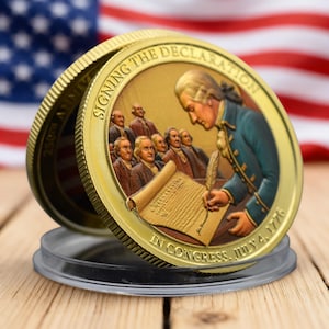 May include: A gold-coloured commemorative coin showing the signing of the Declaration of Independence. The coin features an image of the historical event, with the text "Signing the Declaration" and "In Congress, July 4, 1776". The coin is displayed in a clear plastic case.