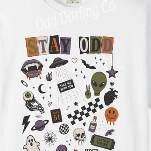 May include: A white t-shirt featuring a graphic print with the text STAY ODD, TAKE ME WITH YOU, AREA 51, BRB, and EARTH IS BORING. The design includes illustrations of a vampire mouth, UFO, alien, skull, ghost, planet, spiderweb, and various symbols.