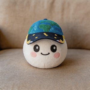 May include: A plush, white, egg-shaped character with a smiling face and pink cheeks. It wears a blue baseball cap decorated with a map of the world. The cap has a dark blue brim with yellow accents and star details. The toy is sitting on a beige surface.