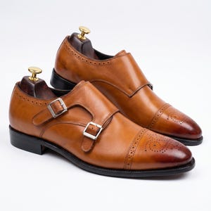 May include: A pair of tan leather double monk strap shoes with silver buckles. The shoes feature a cap toe design with decorative perforations. They are displayed on wooden shoe trees.