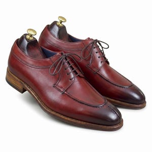 May include: A pair of burgundy leather dress shoes with brown laces and wooden shoe trees. The shoes have a classic design with a cap toe and a subtle contrast in the toe color. The soles are a light brown color.