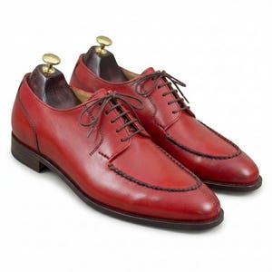 May include: A pair of red leather dress shoes with brown laces. The shoes have a classic design with a stitched border around the toe and a dark brown sole. Shoe trees are inserted inside the shoes.