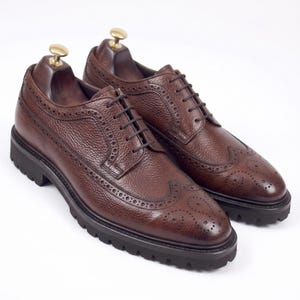 Peut inclure: Une paire de chaussures brogues en cuir marron avec une finition grainée texturée, des détails classiques de type wingtip avec des perforations décoratives, des lacets marron foncé et une épaisse semelle crantée en caoutchouc noir.