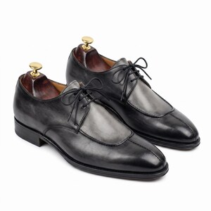 May include: A pair of men's dress shoes in a dark gray and black color scheme. The shoes feature a unique design with a lighter gray panel on the vamp and classic lace-up closure. The shoes are displayed on wooden shoe trees.