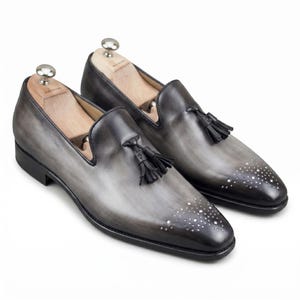 May include: A pair of gray leather loafers with black tassels and silver stud accents on the toe. The shoes have a gradient effect, transitioning from light gray to dark gray. Wooden shoe trees are inserted inside.