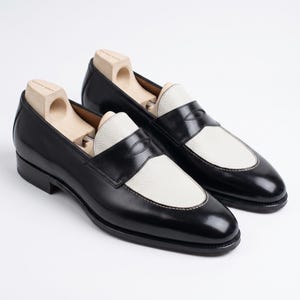 May include: A pair of black and white leather penny loafers with wooden shoe trees inside. The shoes have a classic design with a black upper and a white vamp and saddle. The soles are black, and the shoes are set against a white background.