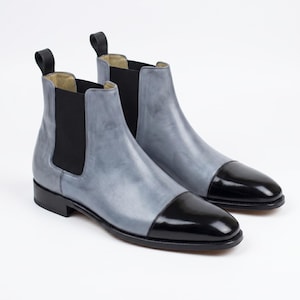 May include: A pair of Chelsea boots. The boots have a two-tone design with a light blue upper and a glossy black toe cap. They feature black elastic side panels and a pull tab at the back.