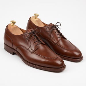 May include: A pair of brown leather dress shoes with a classic design. The shoes feature a rounded toe, lace-up closure, and a polished finish. Shoe trees are inserted inside the shoes. The shoes are displayed on a white background.