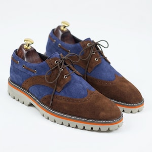 Pode incluir: Um par de sapatos de camurça bicolores em castanho e azul. Os sapatos têm atacadores castanhos, uma sola cinzenta com uma faixa laranja e formas de madeira no interior. O design é do estilo wingtip com perfurações decorativas.