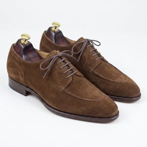 May include: A pair of brown suede dress shoes with a classic design. The shoes feature a rounded toe, a lace-up closure, and a dark brown sole. The shoes are displayed on wooden shoe trees.