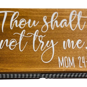 May include: A rectangular wooden sign with the phrase "Thou shalt not try me. MOM 24:7" in white script. The sign has a brown finish and a black and white checkered border.