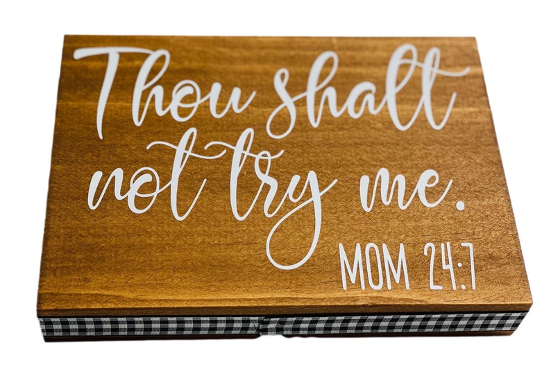 May include: A rectangular wooden sign with the phrase "Thou shalt not try me. MOM 24:7" in white script. The sign has a brown finish and a black and white checkered border.