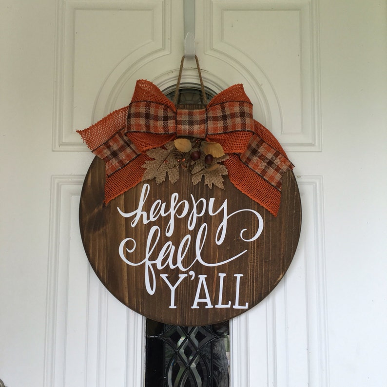 May include: A round wooden door hanger with the text "happy fall y'all" in white lettering. The hanger is decorated with a large orange and brown plaid bow and fall-themed embellishments.