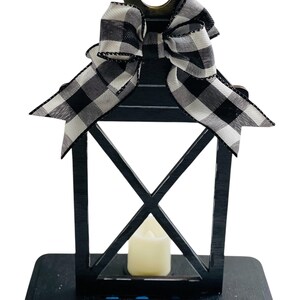 May include: Black wooden lantern with a white and black checkered bow on top. The lantern has a clear glass window and a white candle inside. The lantern is sitting on a black wooden base.