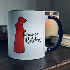 May include: White ceramic mug with a black interior and handle. The mug features a red illustration of a figure in a cloak and the phrase "praise be Bitches" in black script.