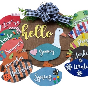 May include: A wooden door hanger with a goose design and the word "hello" in yellow letters. The goose is surrounded by 12 colorful, decorative shapes with words like "Love", "Santa", "Summer", "Pumpkin", "Halloween", "Goosey", "Spring", "Peeps", "Turkey", "Winter", "America", and "Shenanigans".