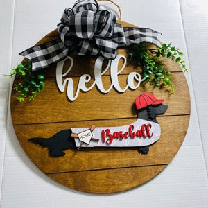 May include: A round wooden door hanger with a brown woodgrain finish. The door hanger features a black and white checkered bow, green foliage, a white "hello" sign, and a black silhouette of a dachshund wearing a red baseball cap and a white "baseball" sign.