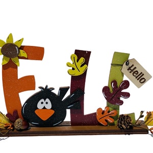 May include: Wooden fall sign with the word "FALL" in orange, red, and green letters. The letters are decorated with glitter, leaves, and a black bird. The sign also includes a small wooden tag that says "Hello".