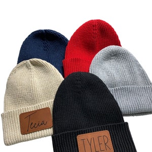 May include: Five knitted beanies in different colors, including red, black, gray, beige, and navy blue. Each beanie has a brown leather patch with a name on it. The names are "Tyler" and "Tecia".