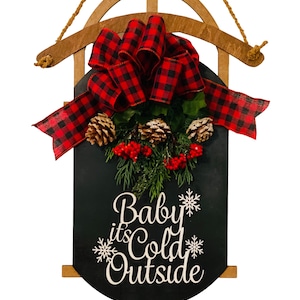 May include: A wooden Christmas sleigh with a black chalkboard sign that reads "Baby it's Cold Outside". The sleigh is decorated with a red and black buffalo plaid bow and pine sprigs with red berries.