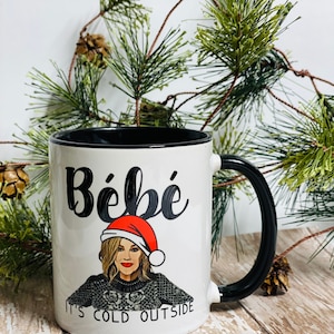 May include: White ceramic mug with a black rim and handle. The mug features a black and white illustration of a woman wearing a red Santa hat and a black and white sweater. The text "Bebé It's Cold Outside" is printed on the mug.