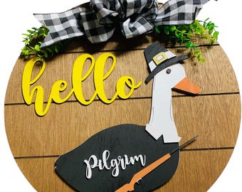 May include: A round wooden door hanger with a black and white checkered bow, green leaves, and a yellow "hello" sign. A black and white pilgrim goose with a hat and a gun is on the bottom of the door hanger. The goose has the word "Pilgrim" written on it.