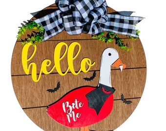 May include: A round wooden door hanger with a Halloween theme. The design features a goose in a vampire costume with the words "Bite Me". The word "hello" is written in yellow. A black and white plaid bow and greenery are at the top.