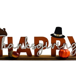 May include: A wooden sign with the words "Happy Thanksgiving" in white lettering. The sign is decorated with a small turkey, a pilgrim hat, and a small pumpkin.