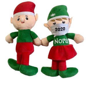 May include: Two plush Christmas elves holding hands. The elf on the left is wearing a red hat and green pants. The elf on the right is wearing a green hat with a snowflake and a white face mask with the text "2020" and "Nora".
