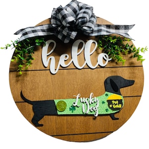 May include: Wooden round sign with a dachshund dog design. The sign features the word "hello" in white script, a black and white checkered bow, and the words "Lucky Dog" with a pot of gold. Greenery accents the top.