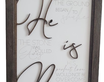May include: Wooden framed sign with the words "He is Risen" in a dark brown, three-dimensional font. The background is white, with additional text in a light gray script.