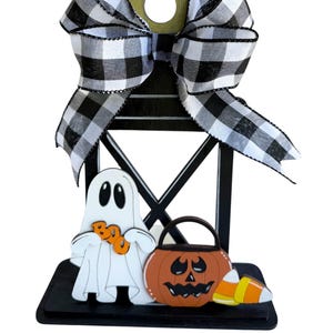 May include: A black wooden Halloween lantern decoration with a large black and white checkered bow. The lantern features a white ghost holding orange "BOO" letters, a jack-o'-lantern bucket, and candy corn.