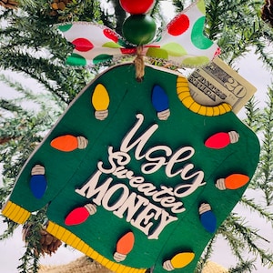 May include: A green wooden Christmas ornament shaped like a sweater with the words "Ugly Sweater Money" and colorful light bulbs. A twenty dollar bill is tucked into the ornament.