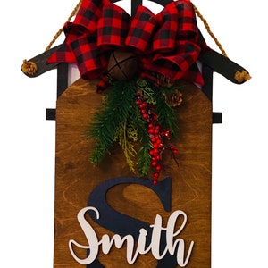 May include: A wooden Christmas sleigh door hanger with a red and black buffalo plaid bow, a bell, and greenery. The name "Smith" is written in white on the sleigh.